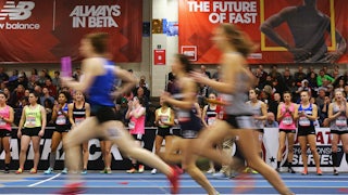 Three young women runners run by, blurred, on an indoor track, as other young runners and spectators look on.