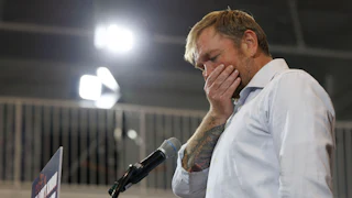 Graham Platner stands at a podium with his hand over his mouth