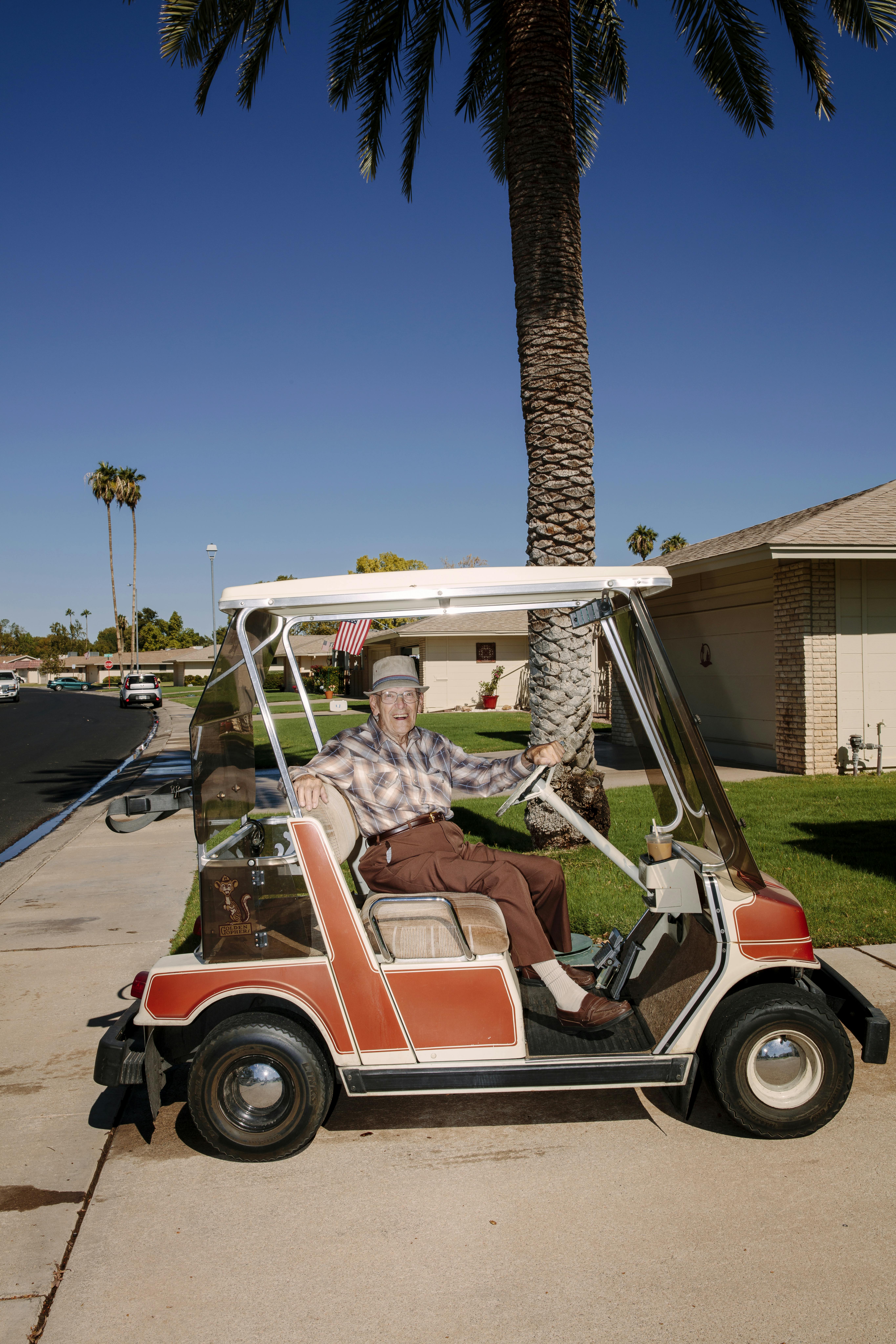 A photograph of an elderly man grinning while seated in a golf cart on a sunny day with palm trees in the background 