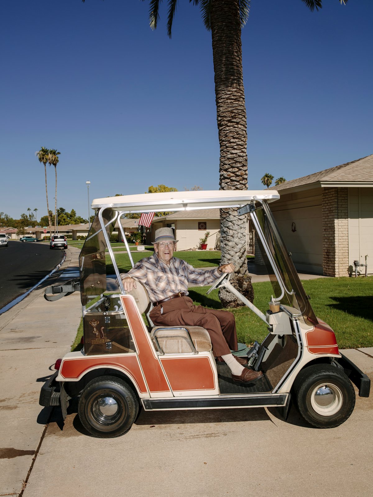 A photograph of an elderly man grinning while seated in a golf cart on a sunny day with palm trees in the background