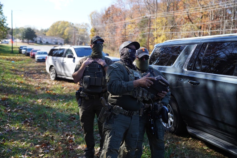 Masked officials stand next to a line of cars.