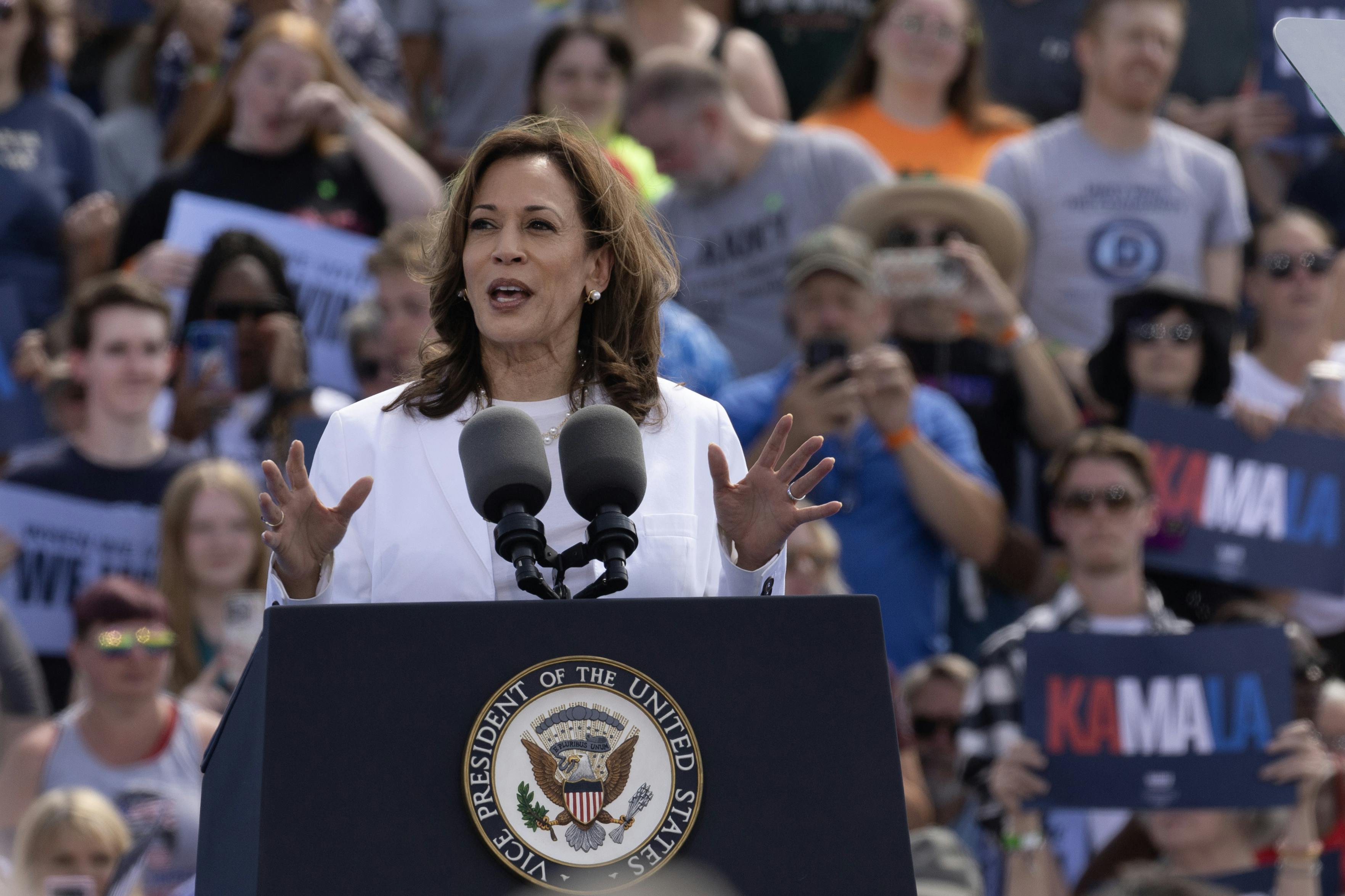 Kamala Harris, dressed in all white, speaks at a podium with supporters behind her.