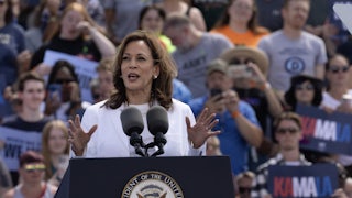 Kamala Harris, dressed in all white, speaks at a podium with supporters behind her.