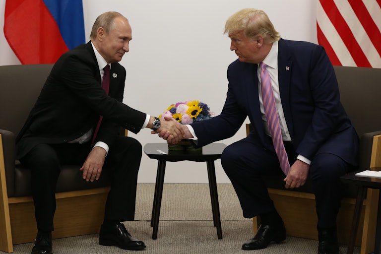 Vladimir Putin and Donald Trump, seated on two chairs, reach over and shake hands.
