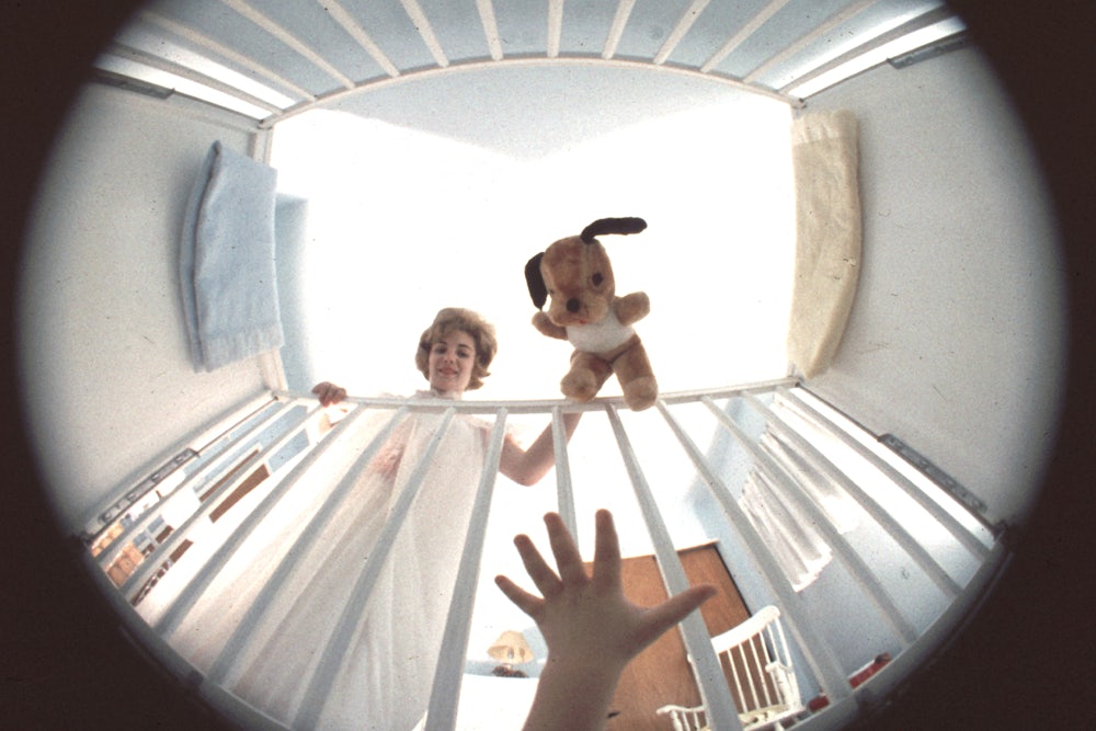A baby's hand reaching up for a stuffed animal, which a woman dangles above the baby in a crib
