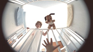 A baby's hand reaching up for a stuffed animal, which a woman dangles above the baby in a crib