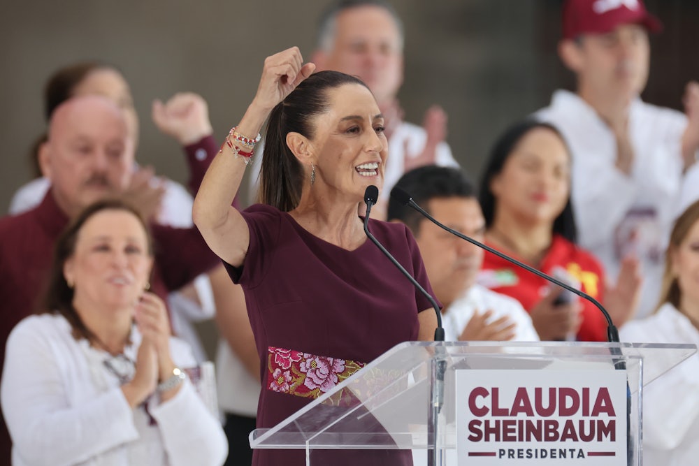 Claudia Sheinbaum raises her fist while standing at a lectern, with people in the background, applauding.