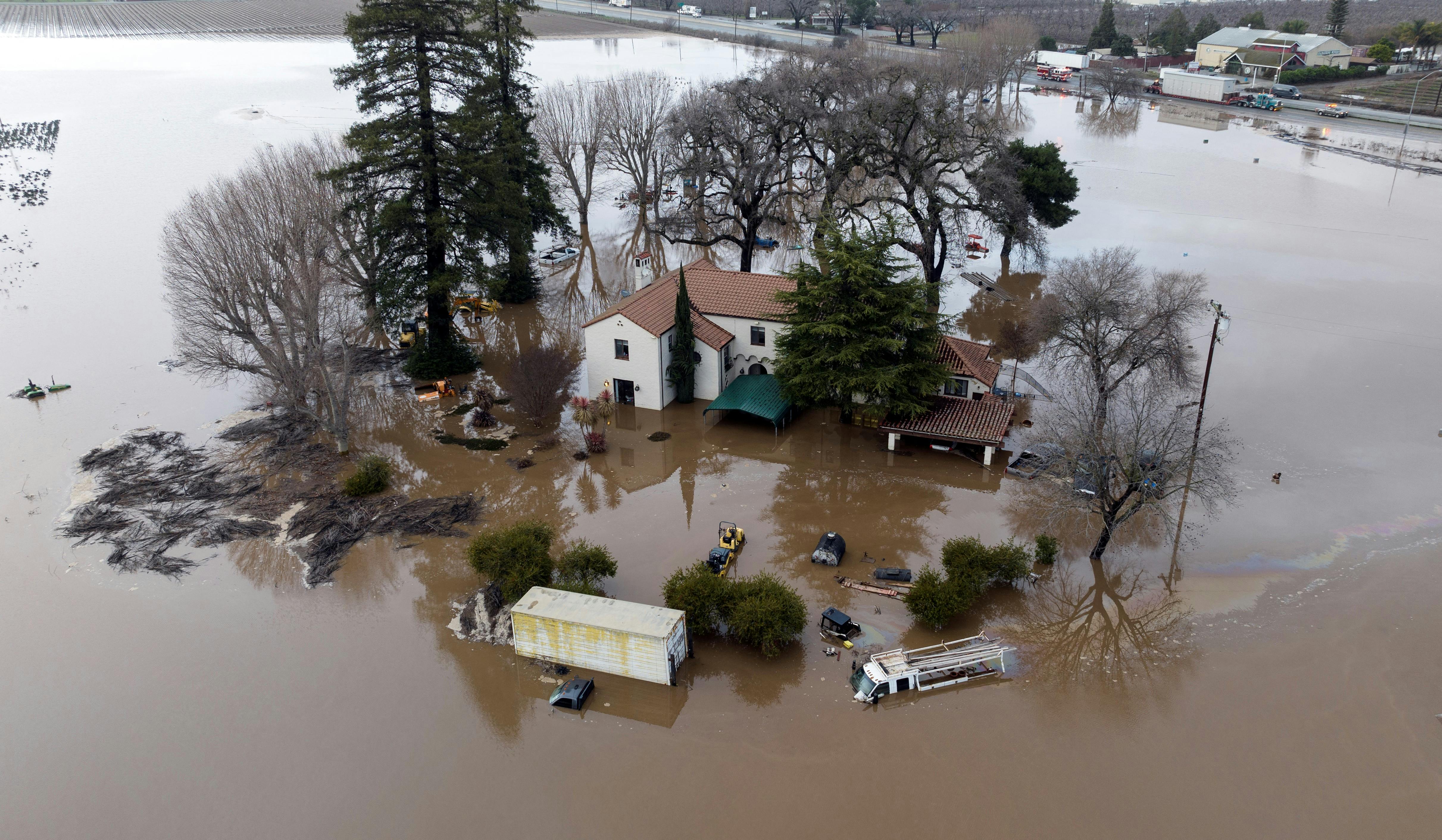 A home, several vehicles, and trees stand flooded with water all around them.