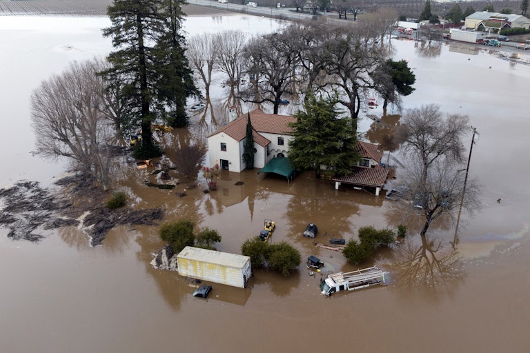 A home, several vehicles, and trees stand flooded with water all around them.