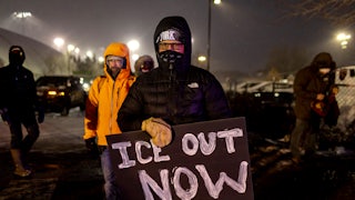 A protester wearing a mask and a large winter coat holds a sign taht reads "ICE OUT NOW."