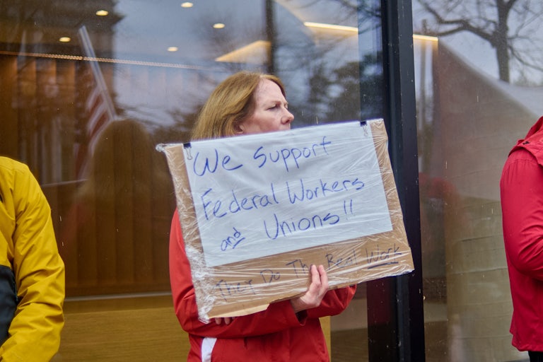 A woman holds a sign reading "We support federal workers and unions!!" while standing outside.