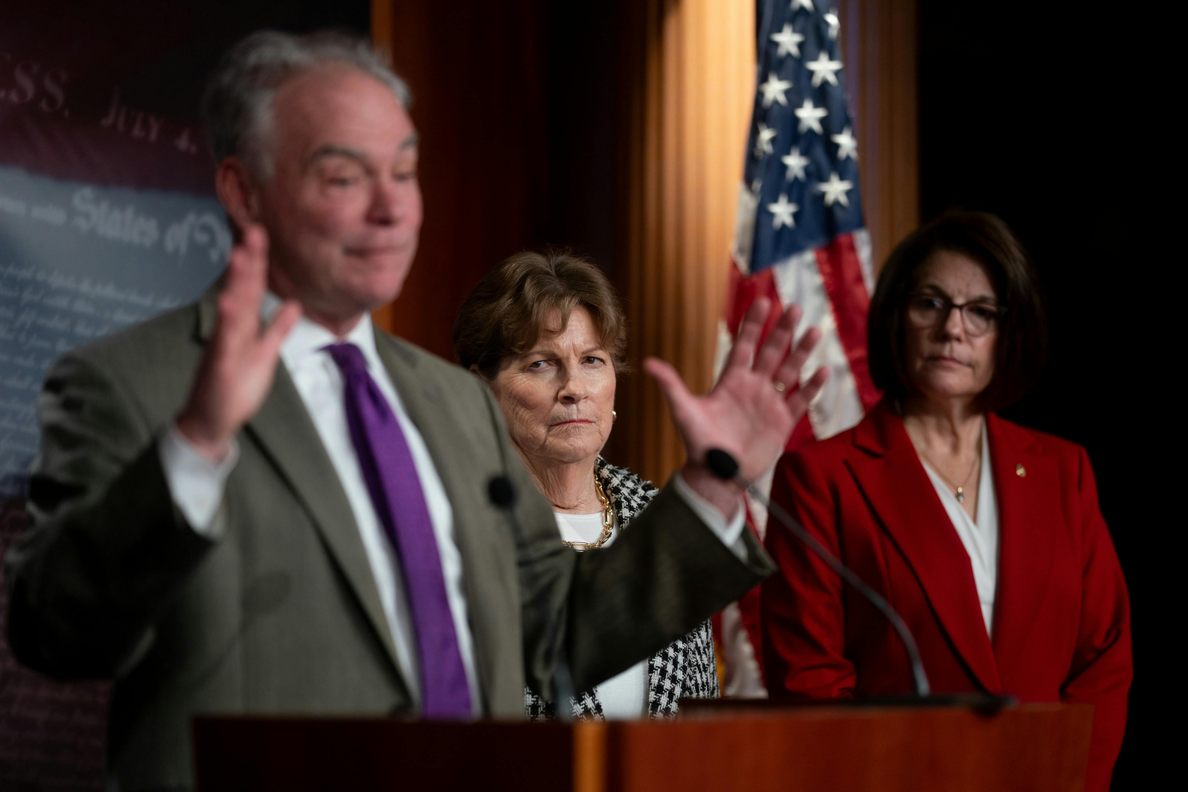 Senator Tim Kaine raises his hands while standing at a podium. Senators Jeanne Shaheen and Catherine Cortez Masto stand behind him