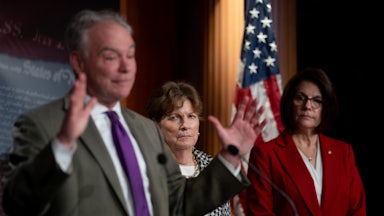 Senator Tim Kaine raises his hands while standing at a podium. Senators Jeanne Shaheen and Catherine Cortez Masto stand behind him