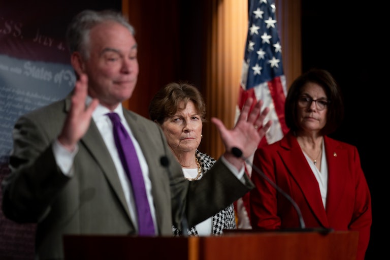 Senator Tim Kaine raises his hands while standing at a podium. Senators Jeanne Shaheen and Catherine Cortez Masto stand behind him
