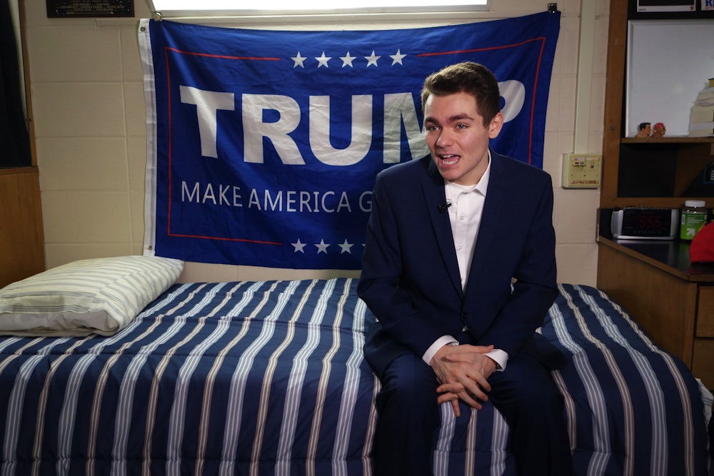 Far-right provocateur and Charlie Kirk-critic Nick Fuentes sits on a bed in front of a blue Trump banner.