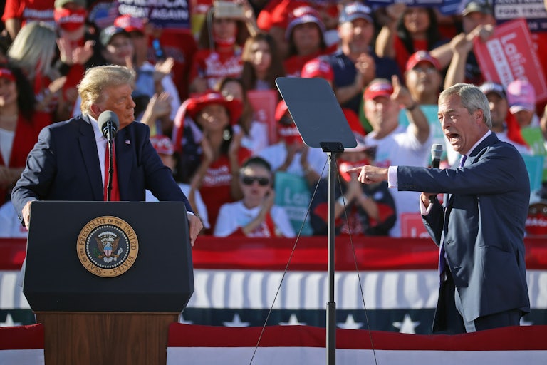 Nigel Farage's mouth is open really, really wide as he speaks into a microphone and points at Donald Trump at a political rally.