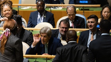 John Kerry leans forward to hear someone speaking to him.