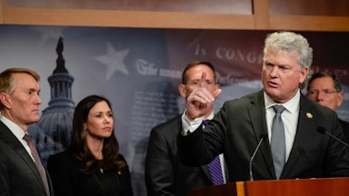 Mike Collins gesticulates while standing at a lectern, with others in suits behind him.