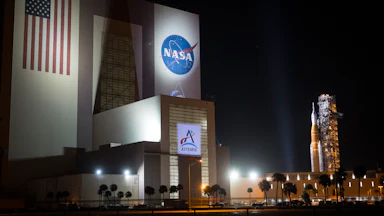 A big building with the NASA logo, an American flag, and Artemis written on it on the left, with a rocket visible in the background on the right.