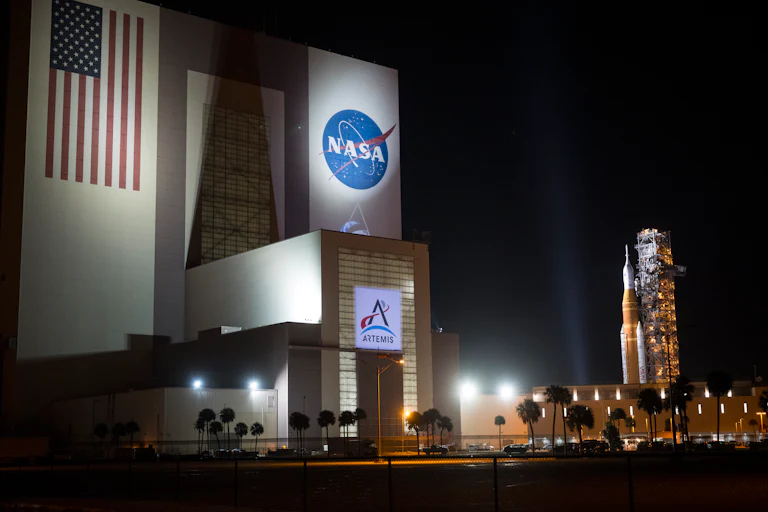 A big building with the NASA logo, an American flag, and Artemis written on it on the left, with a rocket visible in the background on the right.
