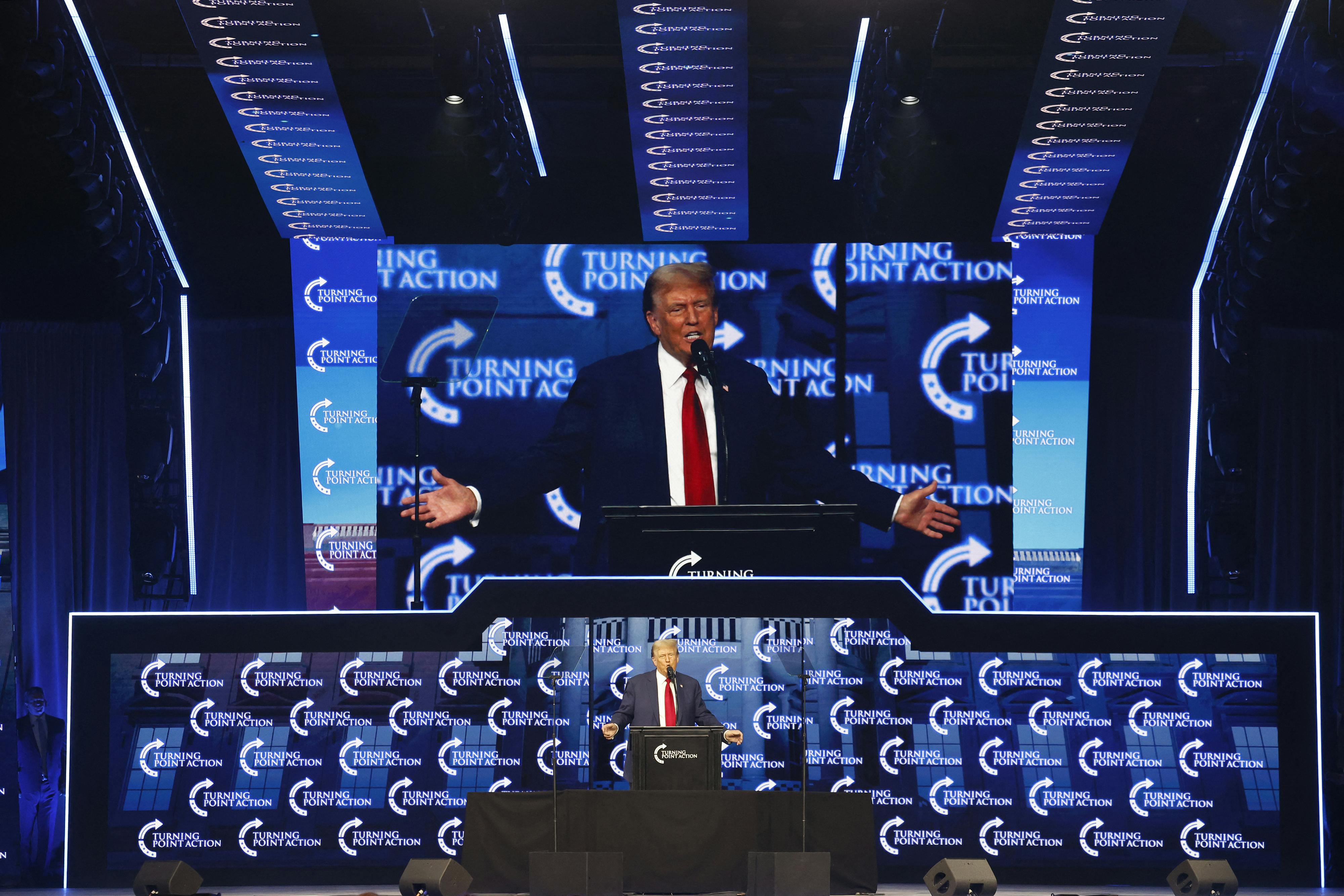 Donald Trump stands at a lectern while his image is projected above him.