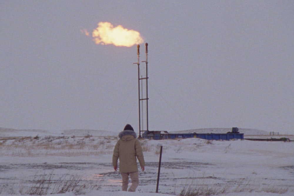 A person walks across a frozen field with a gas or oil flare in the background.