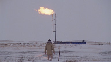 A person walks across a frozen field with a gas or oil flare in the background.
