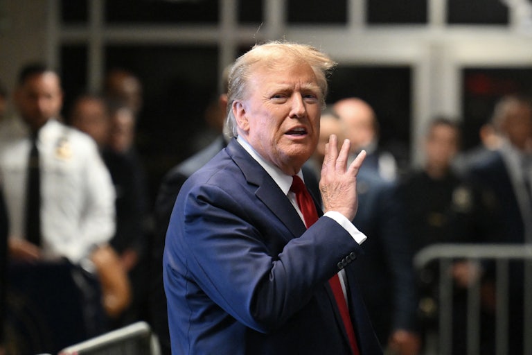 Donald Trump looks agitatd and like he's about to yell something at the camera, hand raised near his mouth. A security guard is in the background, out of focus.