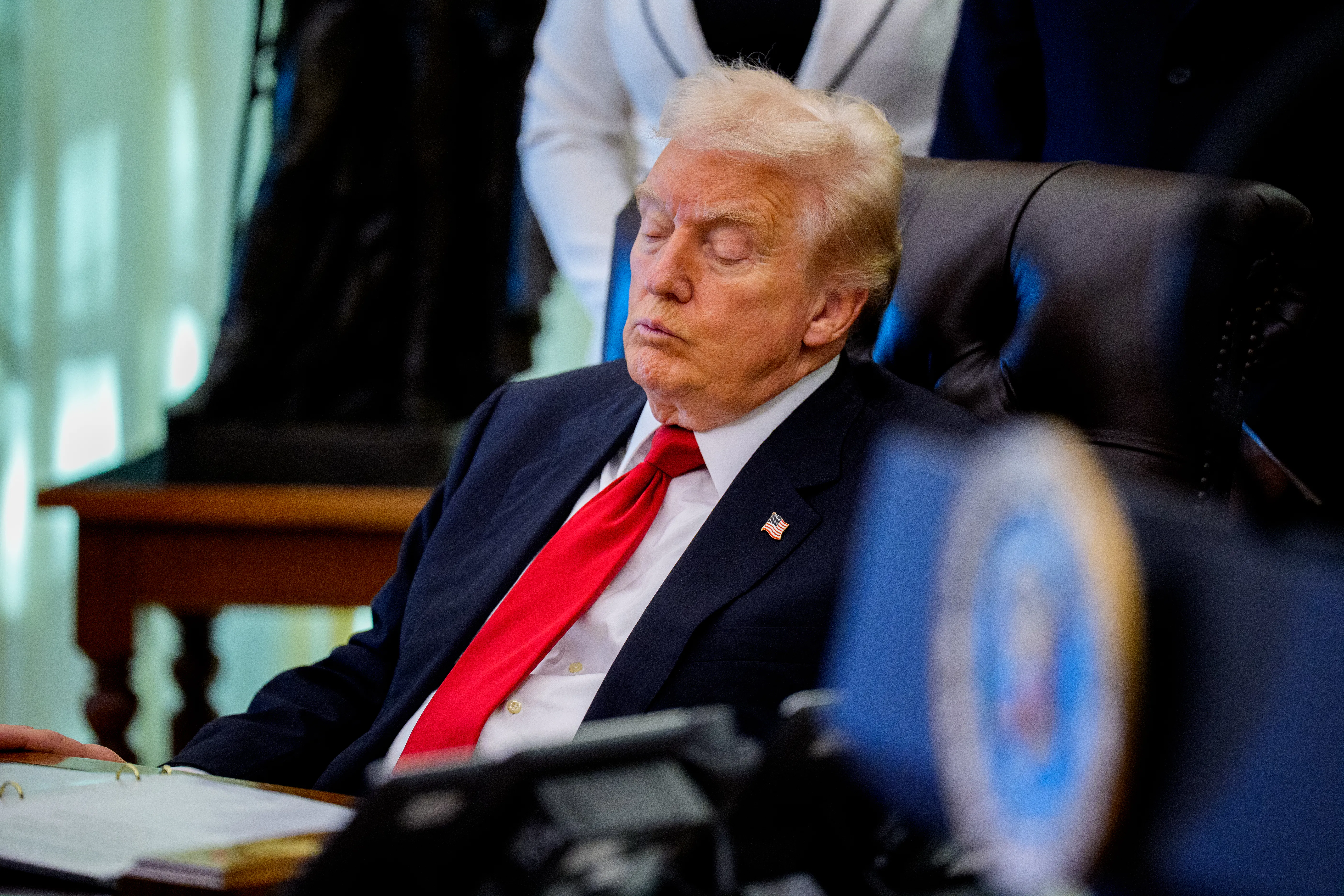 Donald Trump, wearing a blue suit and red tie, sits in side profile on a black leather chair with his eyes closed. In front of him is a desk with an open binder. 