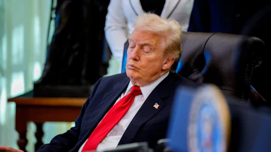 Donald Trump, wearing a blue suit and red tie, sits in side profile on a black leather chair with his eyes closed. In front of him is a desk with an open binder.