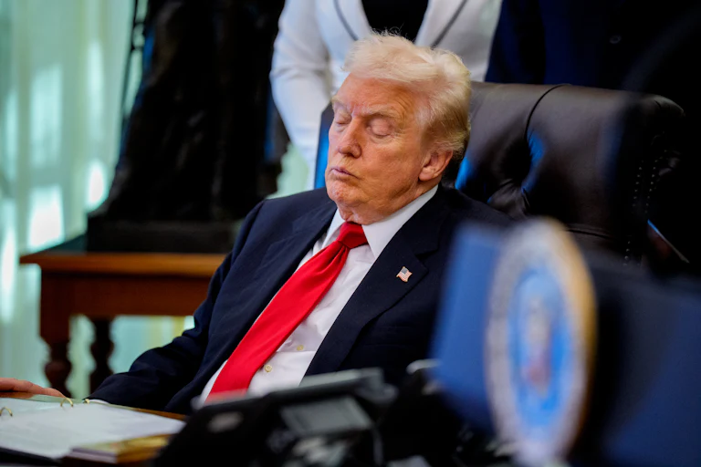 Donald Trump, wearing a blue suit and red tie, sits in side profile on a black leather chair with his eyes closed. In front of him is a desk with an open binder.