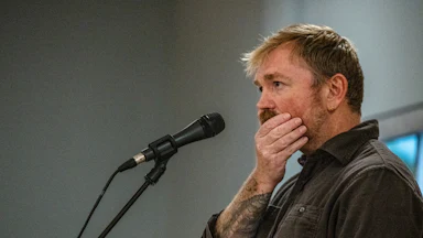 Graham Platner brushes his hands over his jaw while standing before a mic.