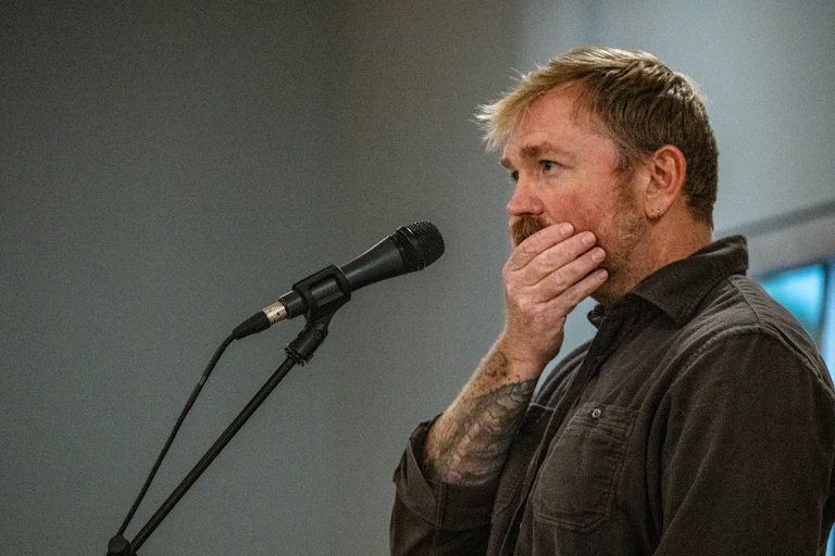 Graham Platner brushes his hands over his jaw while standing before a mic.