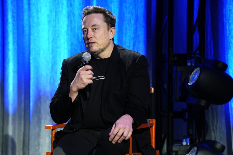 Elon Musk holds a microphone and sits in a chair