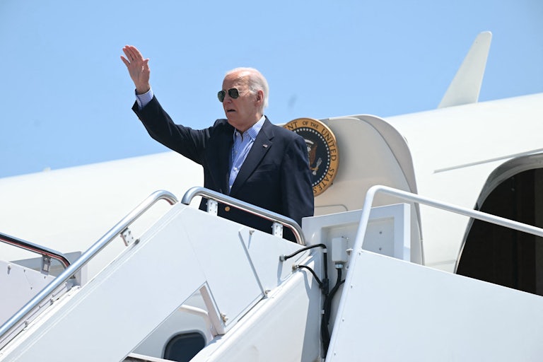 Joe Biden, wearing sunglasses, waves while his mouth hangs open as he stands atop a flight of airplane steps.