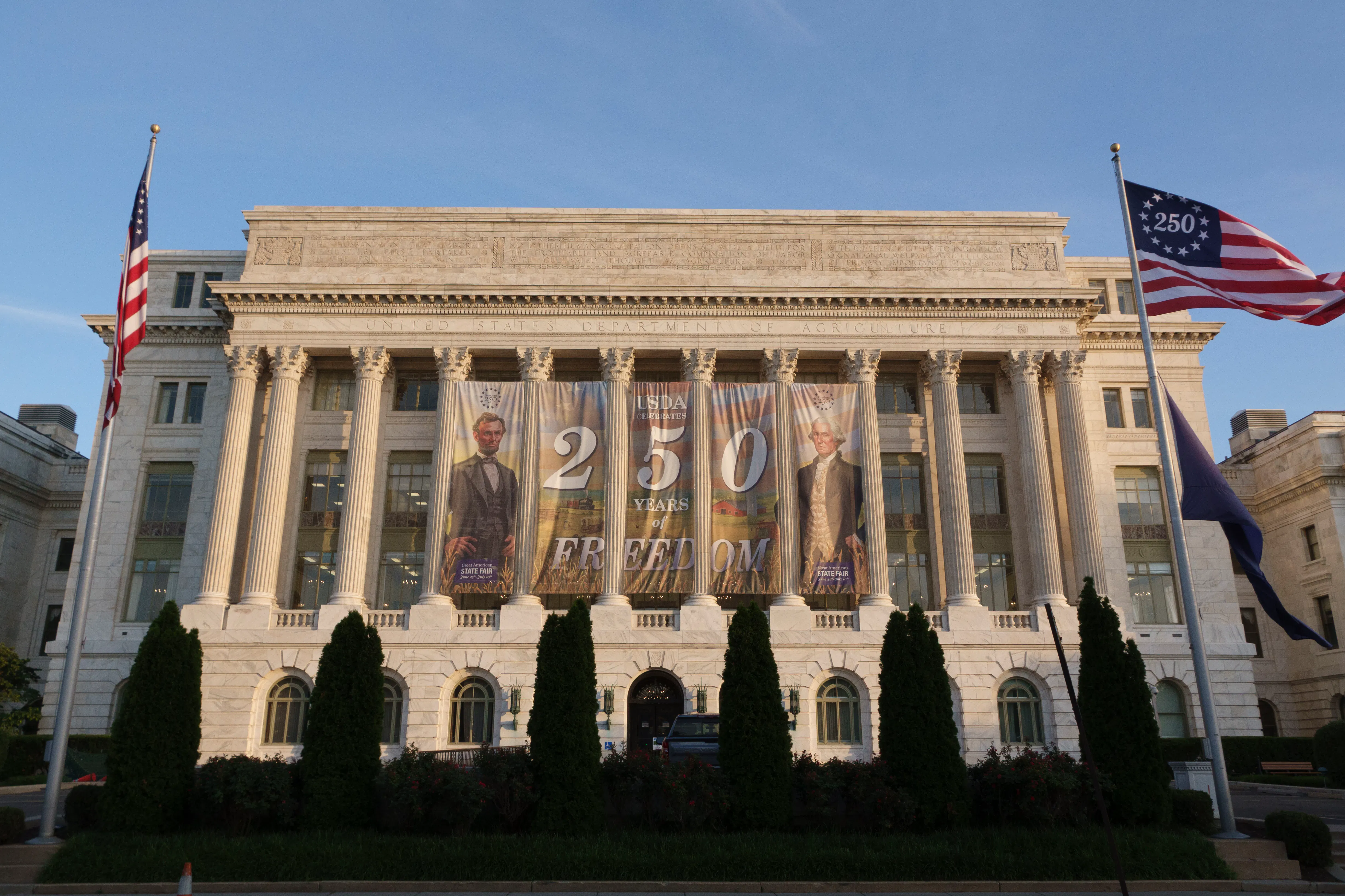 A Freedom 250 banner is hanging behind the columns of the U.S. Department of Agriculture building in Washington, D.C. Two American flags are visible on either side of the building on flagpoles, with "America 250" visible on one of them instead of the normal 50 stars. 