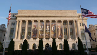 A Freedom 250 banner is hanging behind the columns of the U.S. Department of Agriculture building in Washington, D.C. Two American flags are visible on either side of the building on flagpoles, with "America 250" visible on one of them instead of the normal 50 stars.