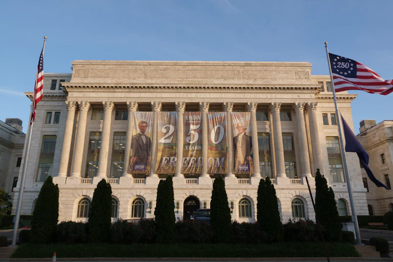 A Freedom 250 banner is hanging behind the columns of the U.S. Department of Agriculture building in Washington, D.C. Two American flags are visible on either side of the building on flagpoles, with "America 250" visible on one of them instead of the normal 50 stars.