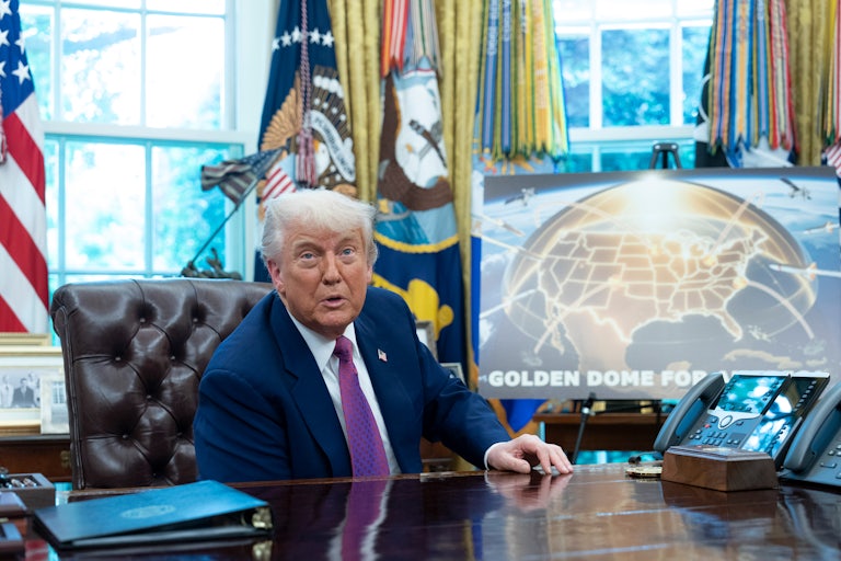 Donald Trump sits at his desk in the Oval Office in front of a poster for the Golden Dome