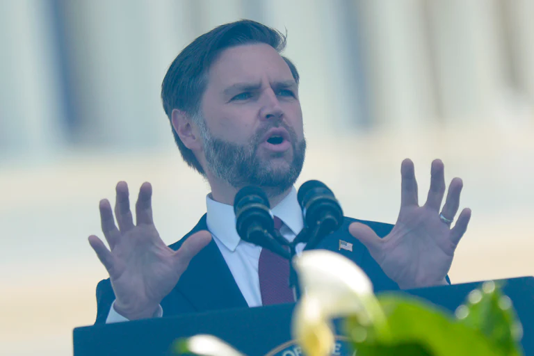 Vice President JD Vance gestures while speaking at a podium behind bulletproof glass