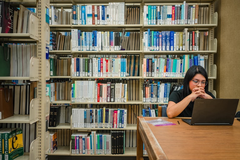 A student sits at a table in the library with her chin resting on her hands, reading the laptop in front of her. A row of bookcases is in the background.