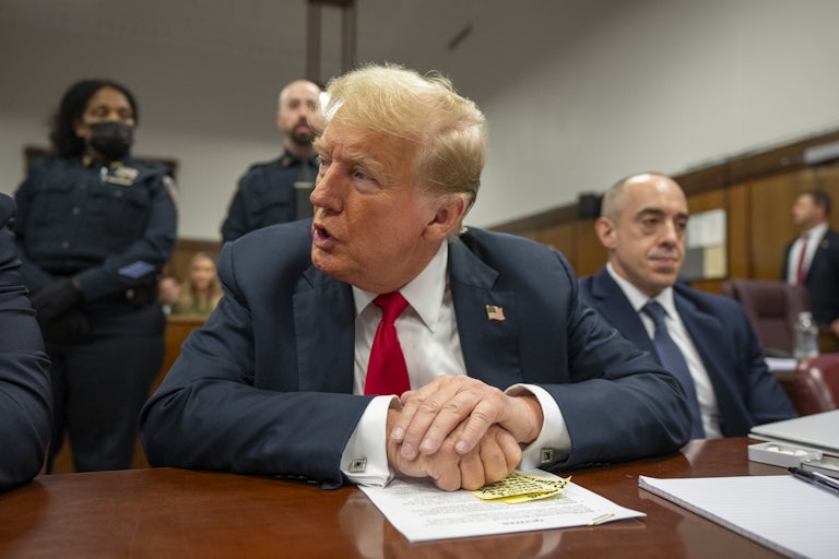 Donald Trump in the courtroom rests his hands on a stack of papers and yellow sticky note. His laywer beside him stares off into the distance.