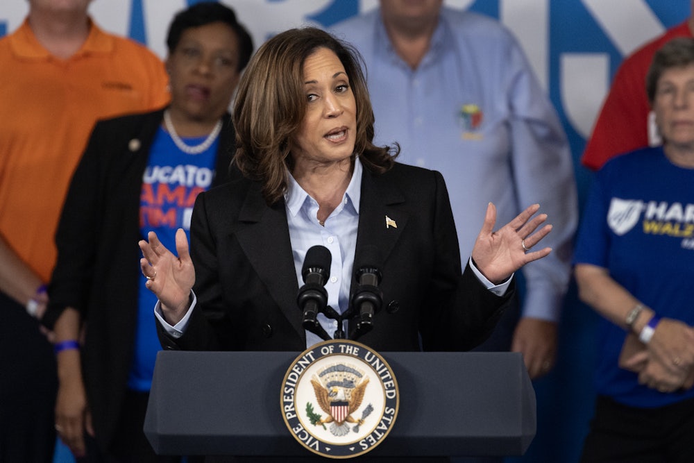 Kamala Harris holds her hands out while speaking at a podium, with other people standing behind her.