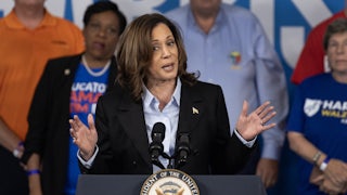 Kamala Harris holds her hands out while speaking at a podium, with other people standing behind her.