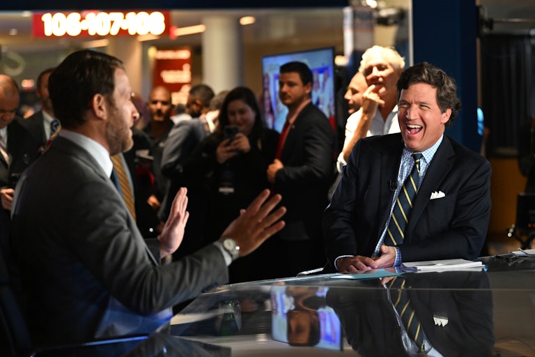 Tucker Carlson laughs while doing an interview with someone seated beside him