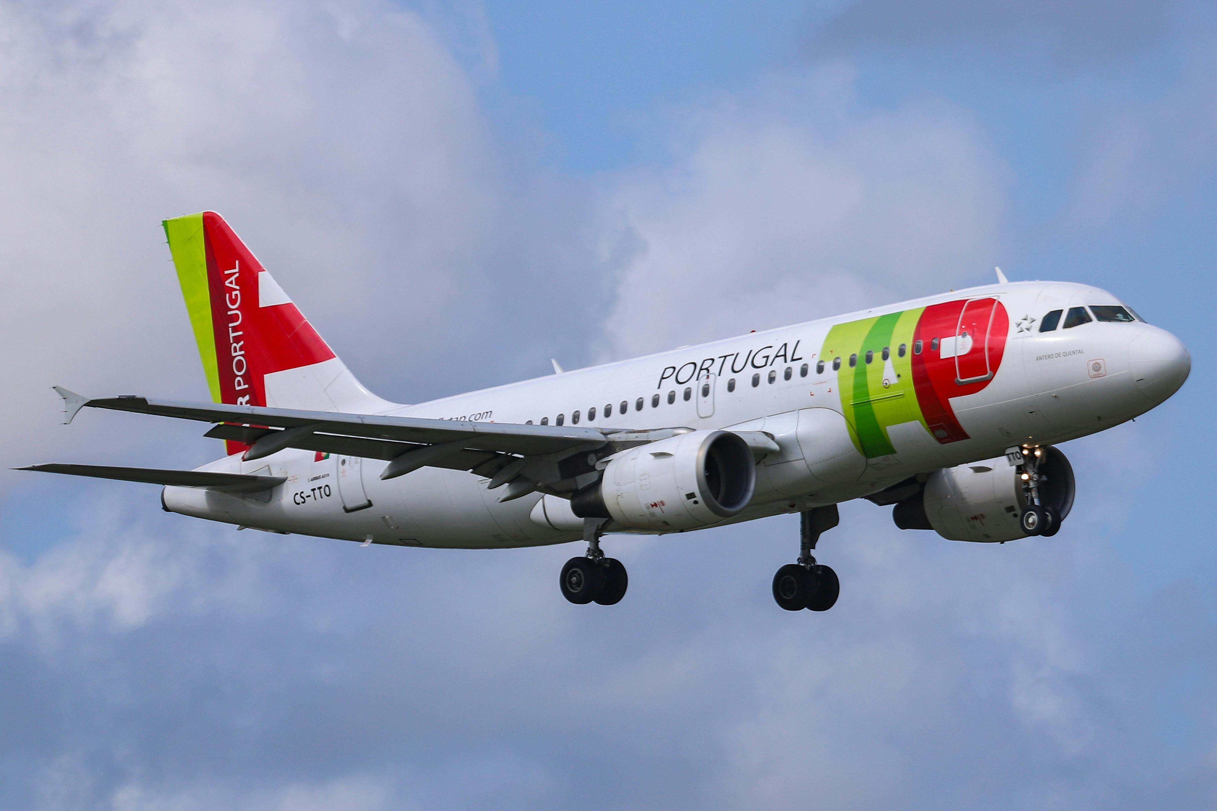 A plane with TAP Air Portugal markings on the side is seen in the air, against a blue sky with clouds.