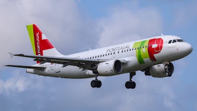 A plane with TAP Air Portugal markings on the side is seen in the air, against a blue sky with clouds.