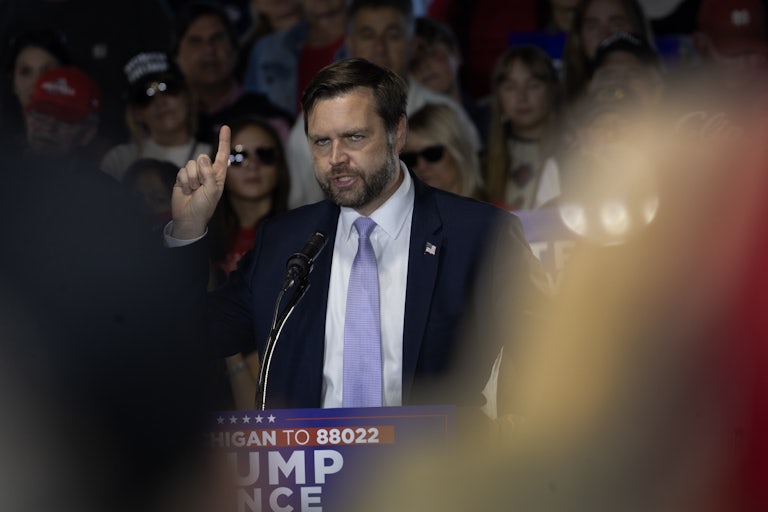 a man wearing a purple tie holds up one finger as he speaks behind a lectern