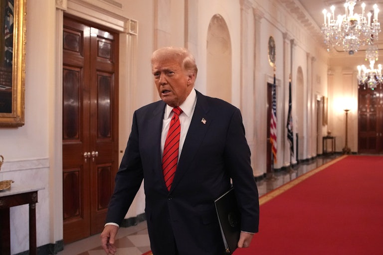 Donald Trump walks down a red carpet in the White House while holding what is presumably an executive order in his left hand.