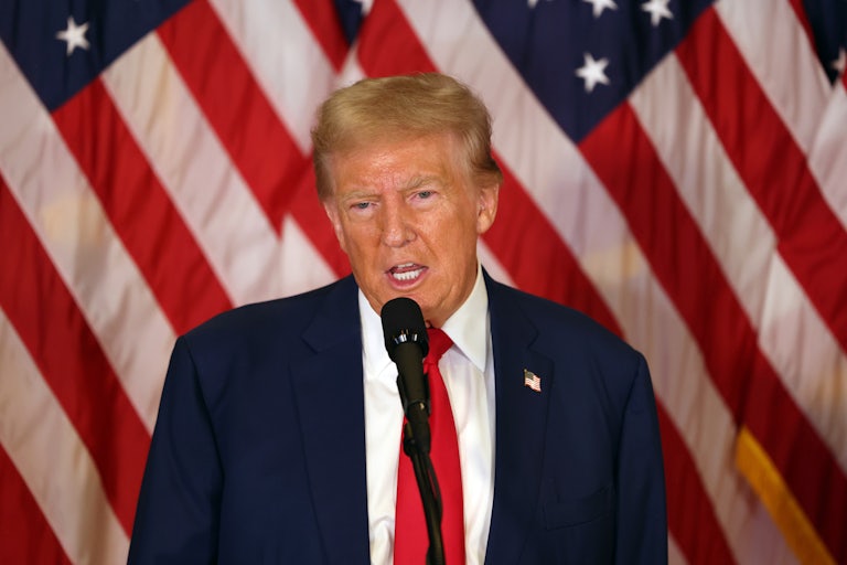 Donald Trump speaks at a microphone. U.S. flags are behind him.